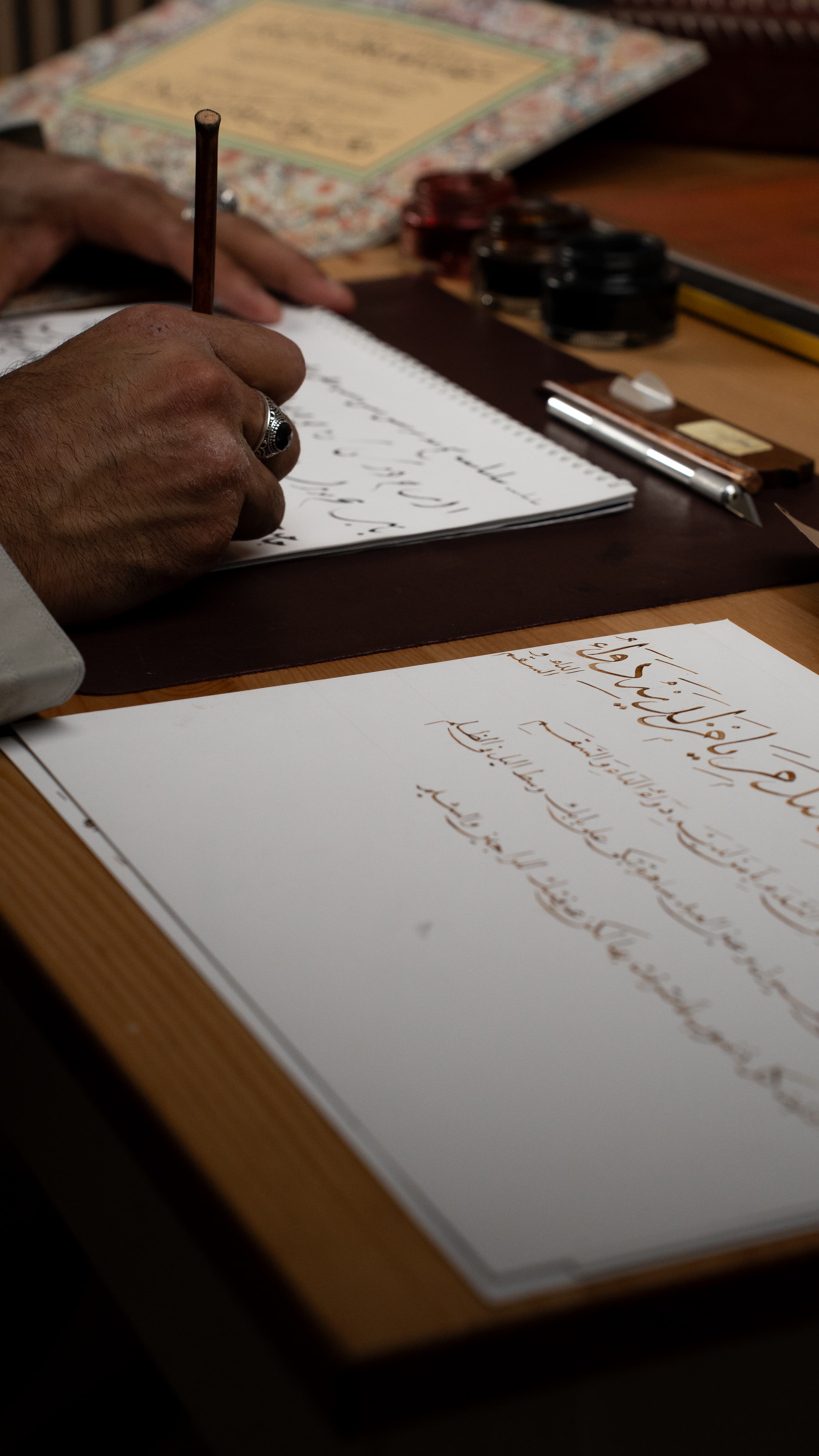 A calligrapher's hand writing Arabic script with a qalam, beside a gold Thuluth panel and ink wells